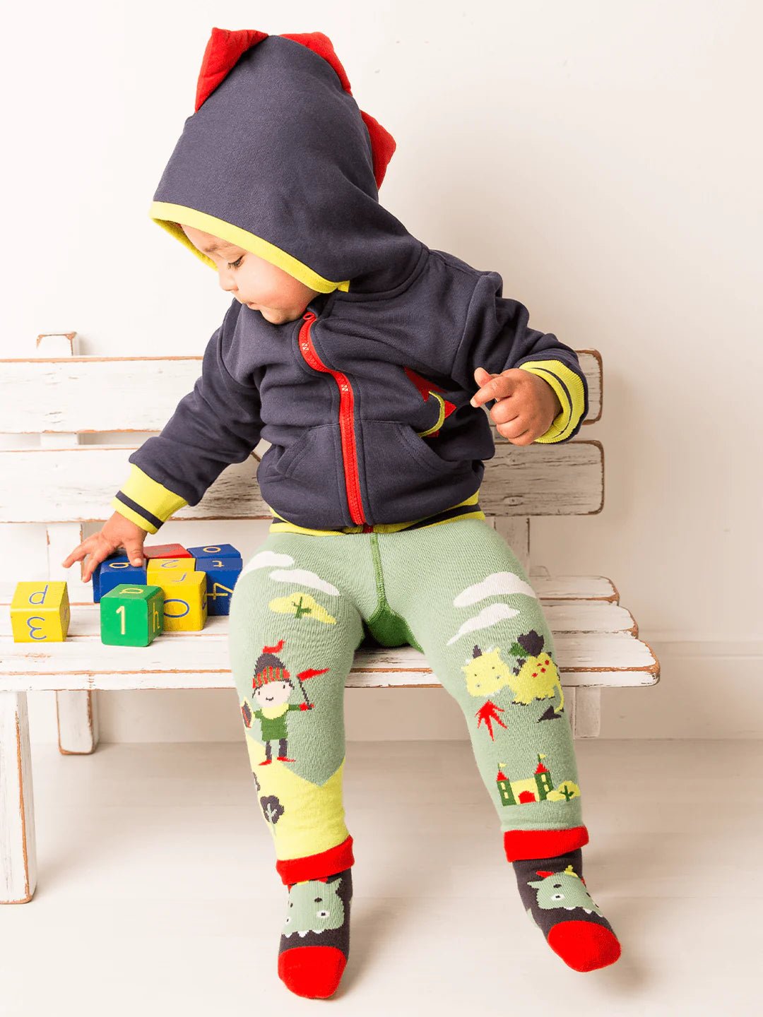 Child wearing a dinosaur-themed outfit with a hoodie and leggings, sitting on a wooden bench.