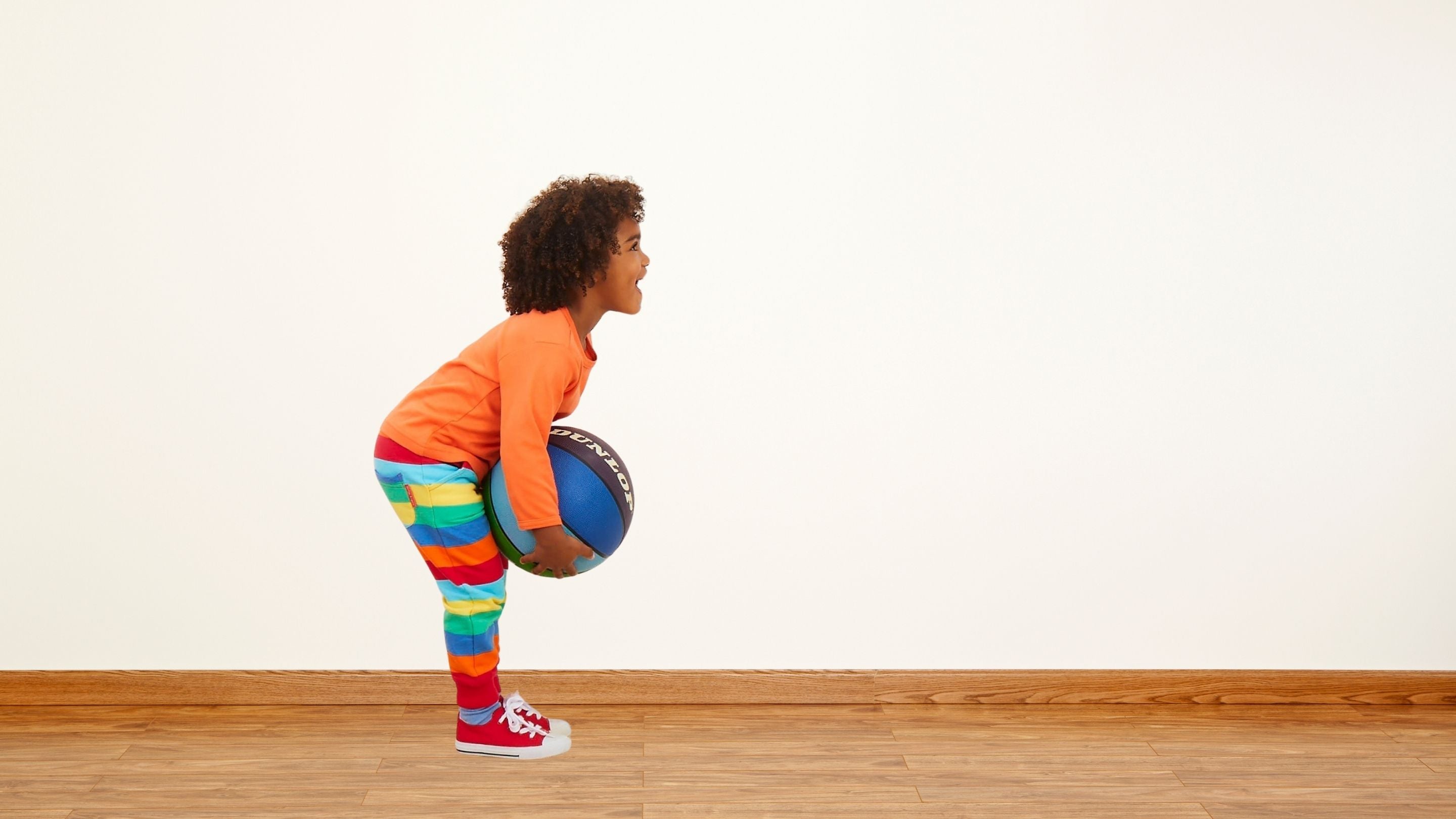 Child in colorful outfit playing with a ball on a wooden floor.