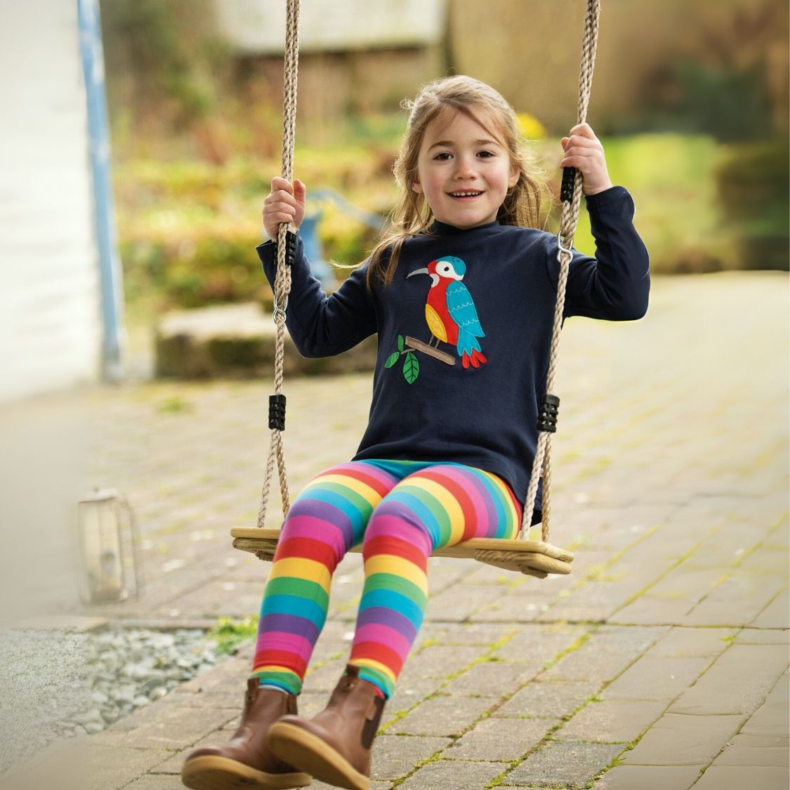 Child on a swing wearing a dark blue sweater with a colorful bird design and rainbow striped leggings.