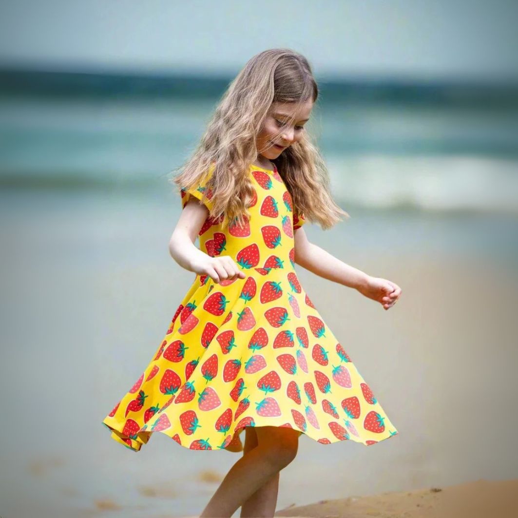 Young girl in a yellow dress with strawberry pattern standing on a beach.