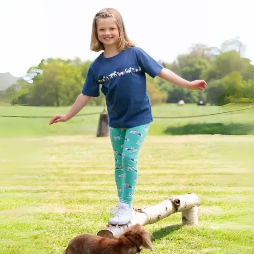 Girl in blue shirt and green leggings standing on a log with a dog in a park