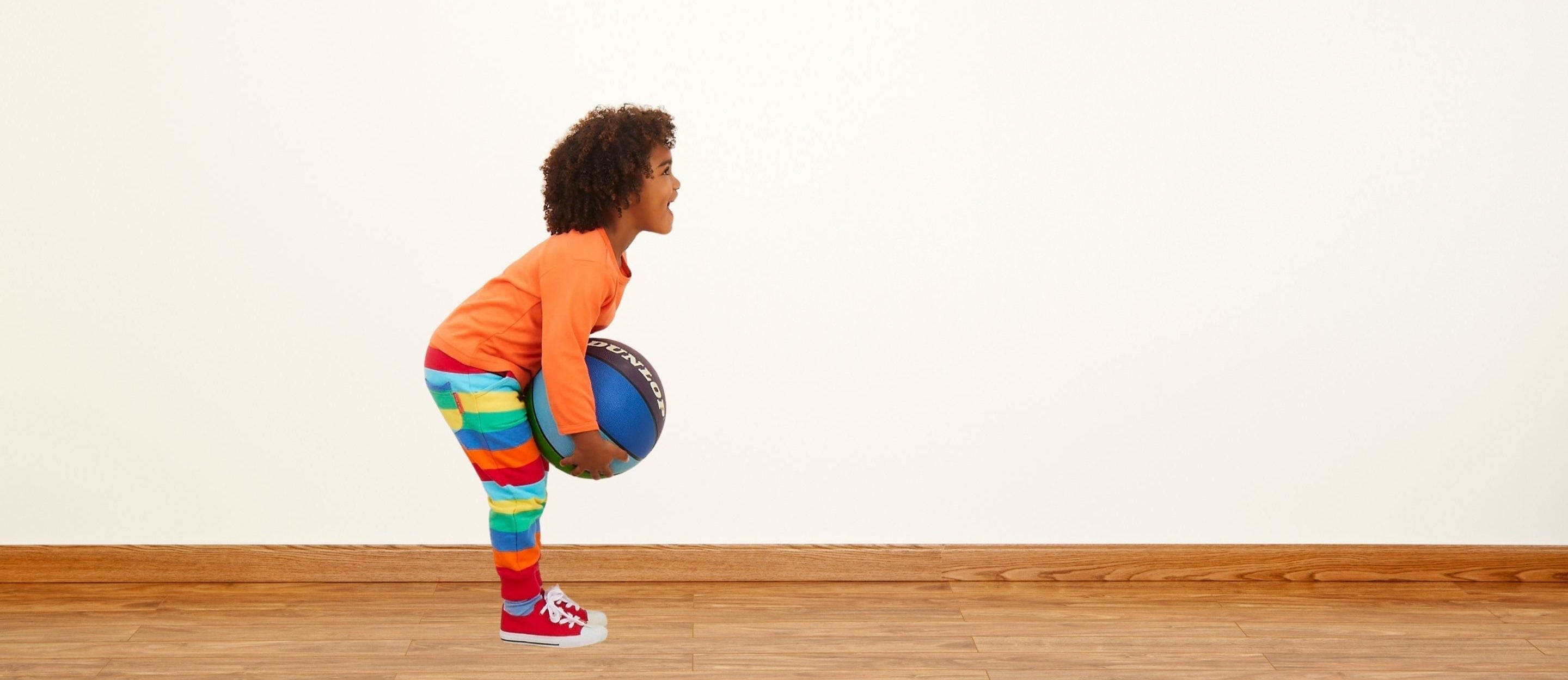 Child holding a basketball in a room with a white wall and wooden floor