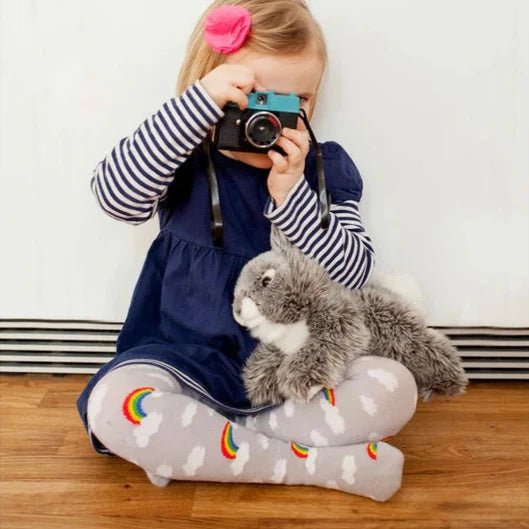 Child holding a camera with a stuffed animal, sitting on a wooden floor against a white wall.