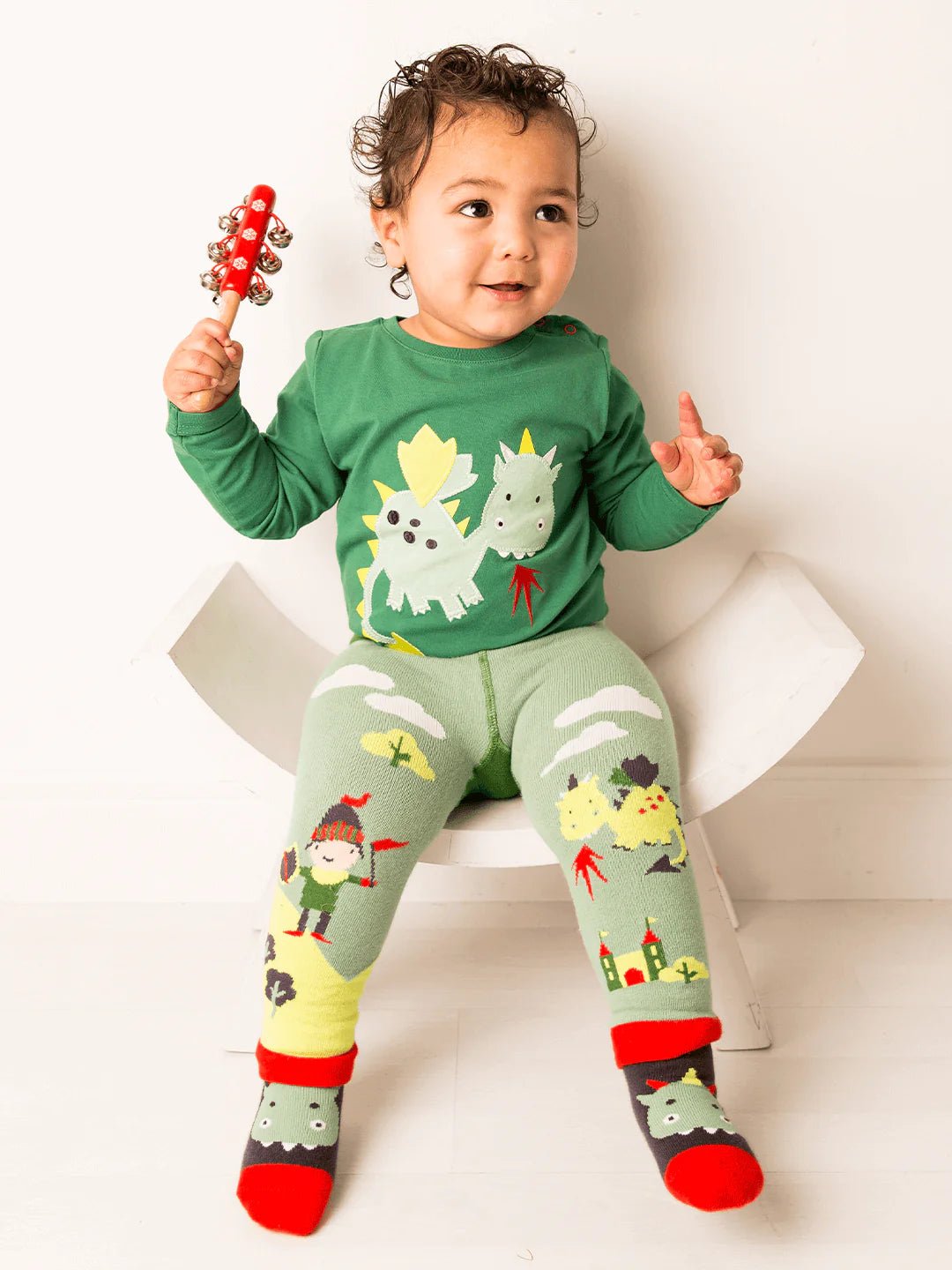 Child wearing a green long-sleeve shirt and matching leggings with cartoon designs, sitting on a white surface.