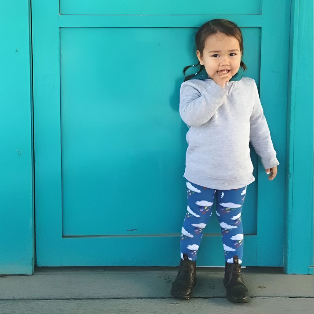 Child wearing a gray sweatshirt and blue leggings with white patterns, standing in front of a teal door.