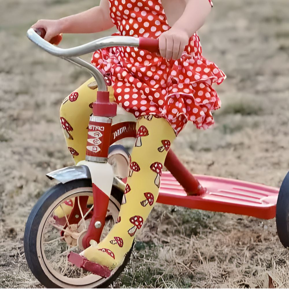 Child riding a tricycle with red and white polka dot dress and yellow leggings with red mushroom pattern.