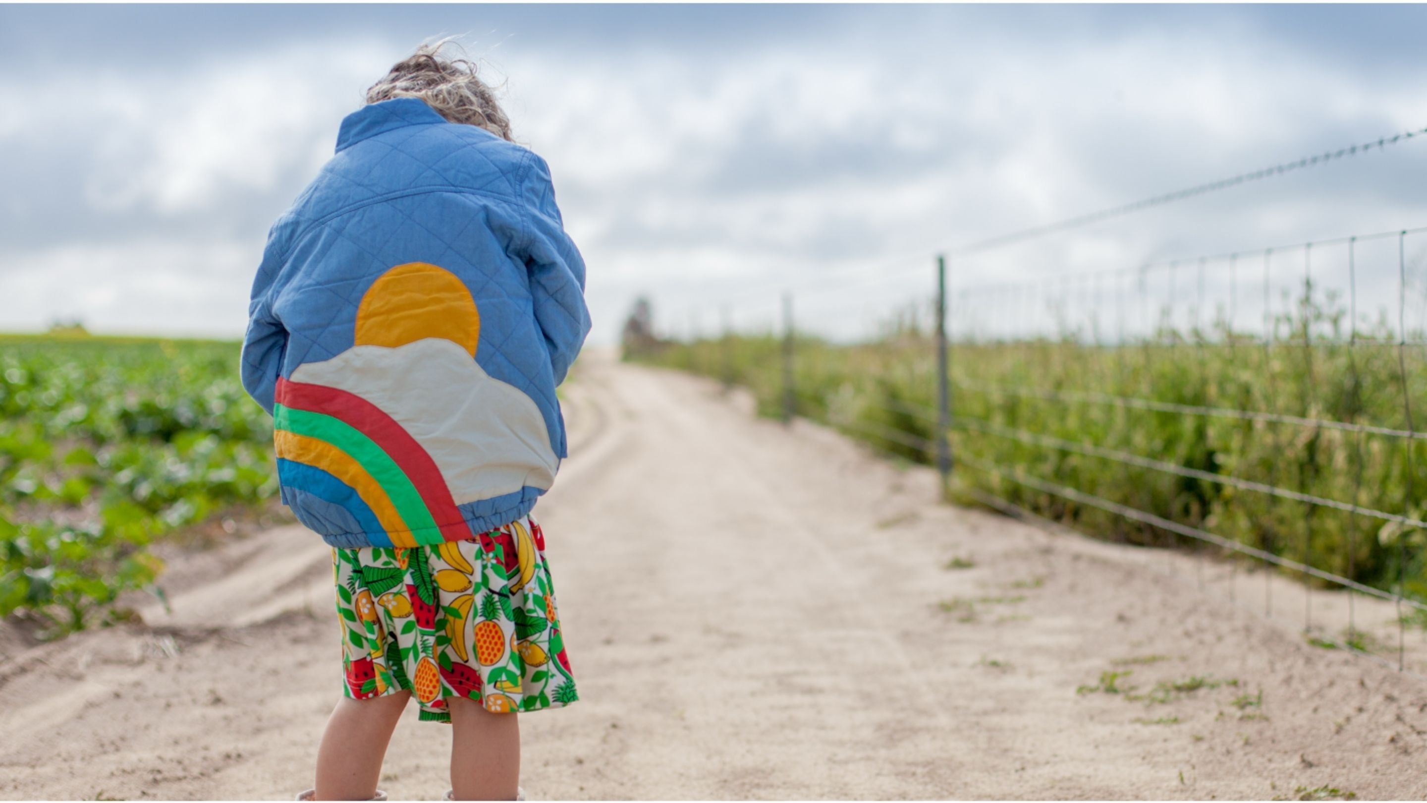 Child wearing blue jacket with sun and rainbow design and fruit print skirt on dirt road by wire fence