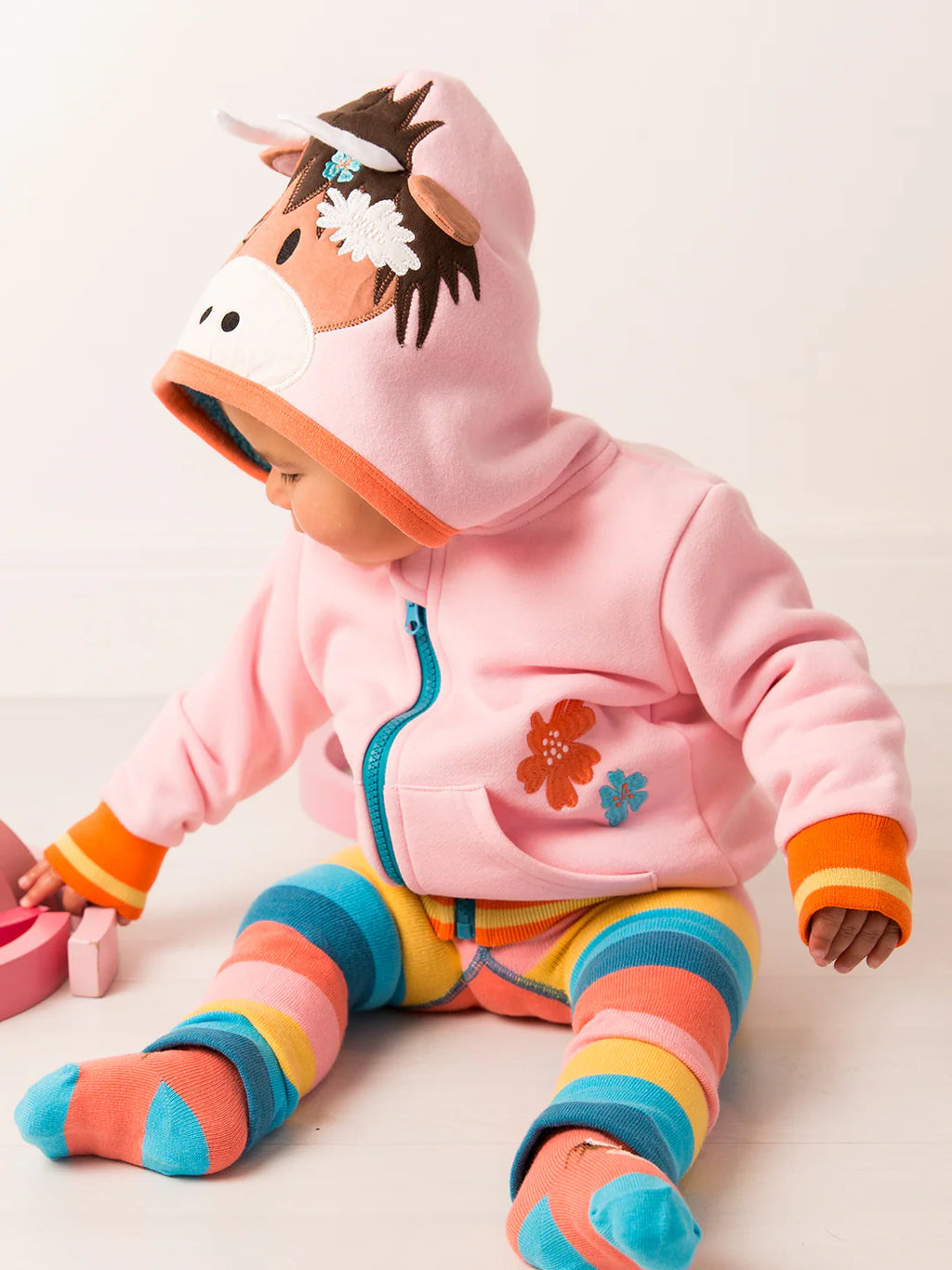 Child wearing a pink hoodie with a horse design and colorful striped pants on a light background
