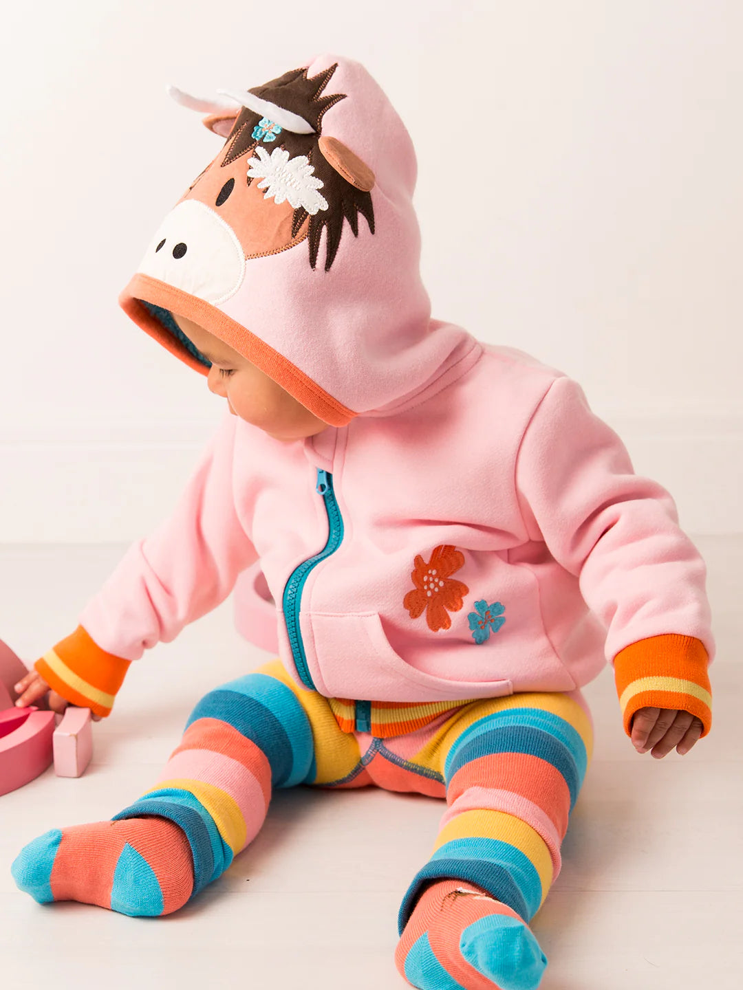 Child wearing a pink hoodie with a horse design and colorful striped pants on a light background