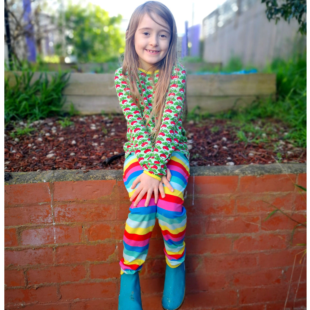 Child wearing a colorful outfit with a patterned top and rainbow striped pants, standing against a brick wall outdoors.