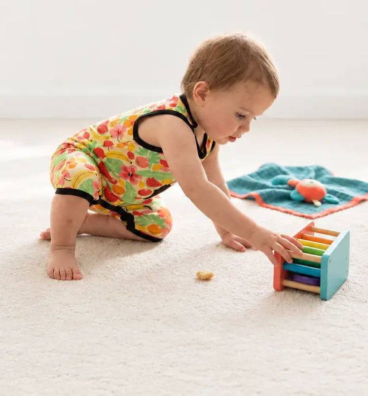 A baby wearing a colorful floral print sleeveless romper crawls on a white carpet, reaching for a small wooden toy. A teal soft toy is visible in the background.