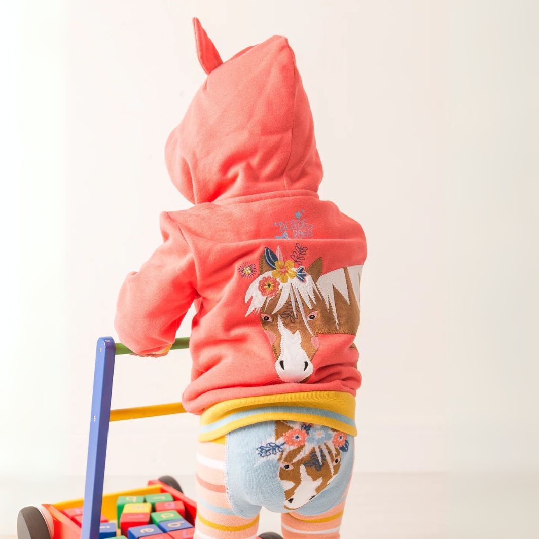 Child wearing a pink hoodie with a horse design, standing next to a toy.