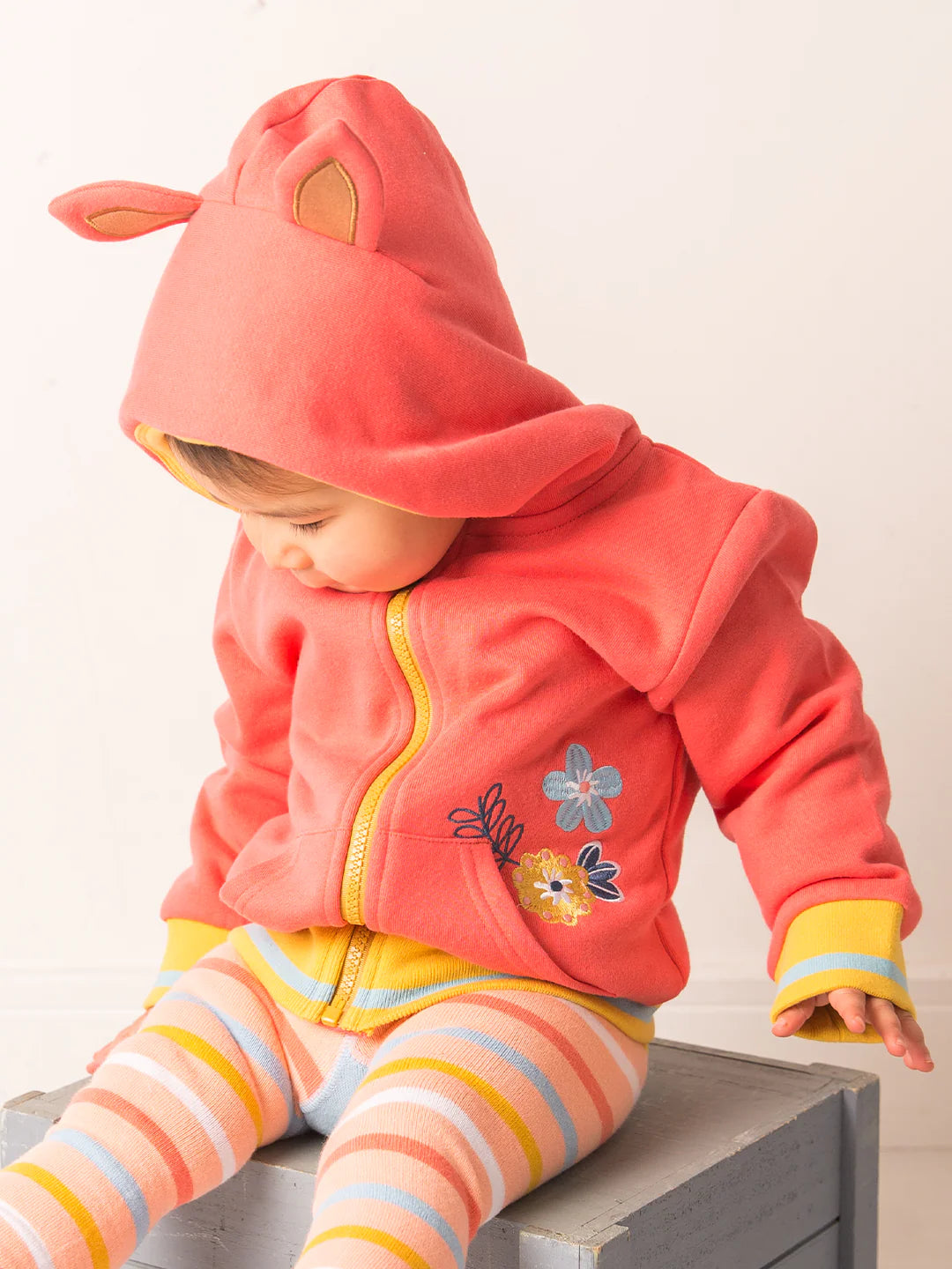 Child wearing a pink hooded jacket with floral designs and striped pants on a light background