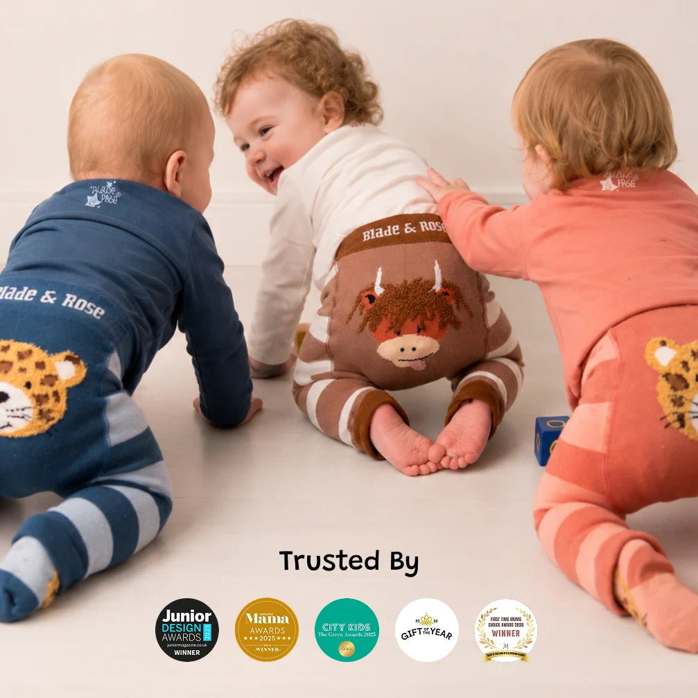 Three babies crawling on a light floor wearing matching 'Blade & Rose' brand onesies with animal and cow graphics. The left baby wears a blue leopard print one-piece, the center baby wears a white and brown striped one-piece with a cow on the back, and the right baby wears a coral pink leopard print one-piece.