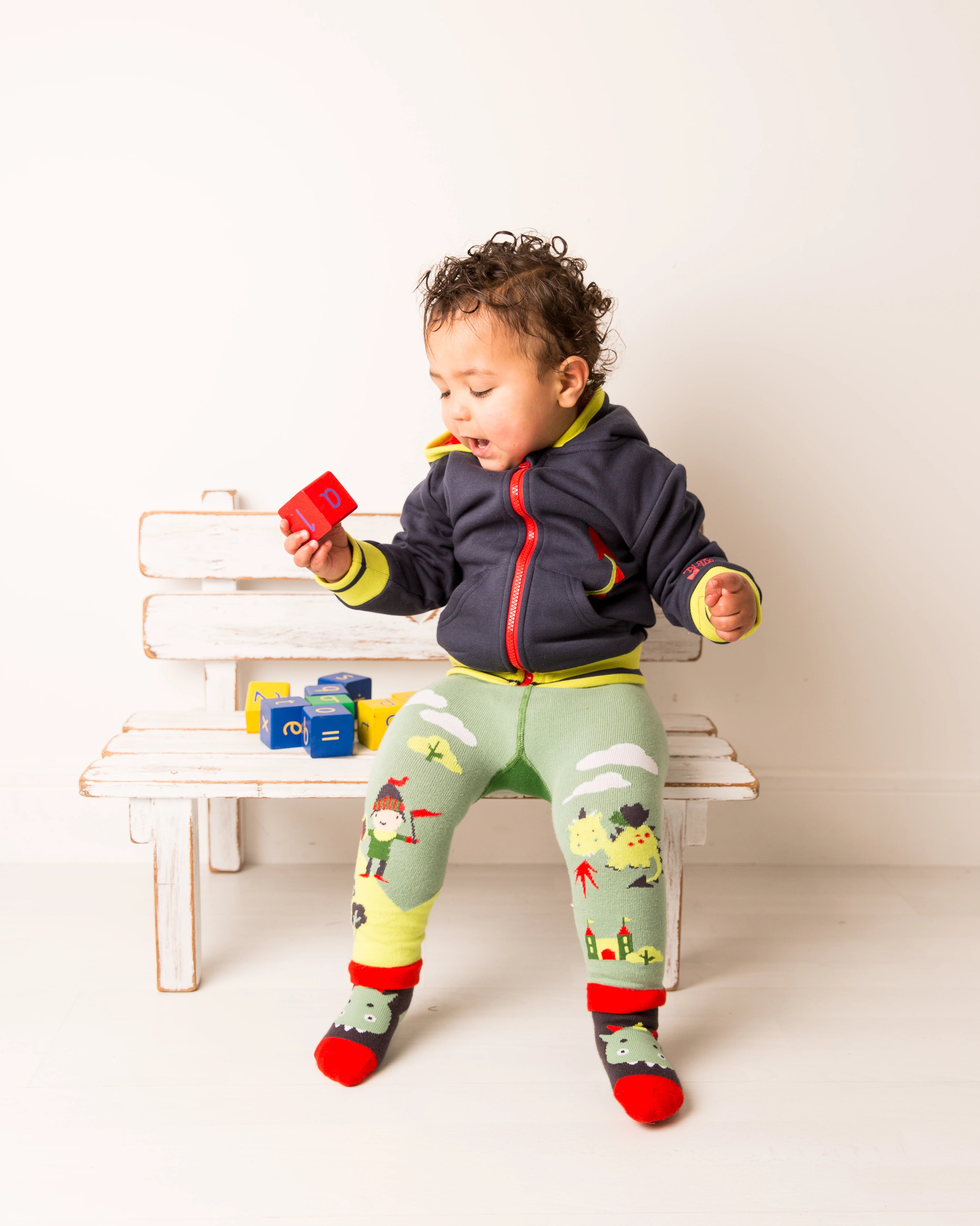 Child wearing a colorful outfit with a jacket, leggings, and socks, sitting on a white bench.