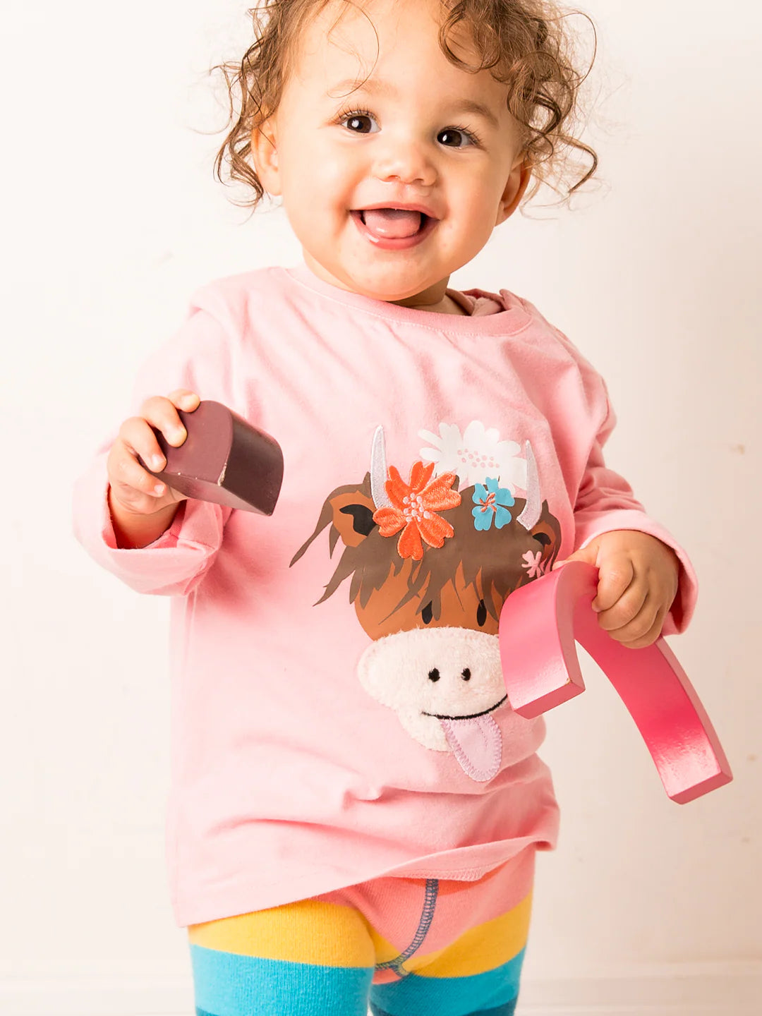 Child wearing a pink shirt with a horse design, holding a brown object and pink ribbon.
