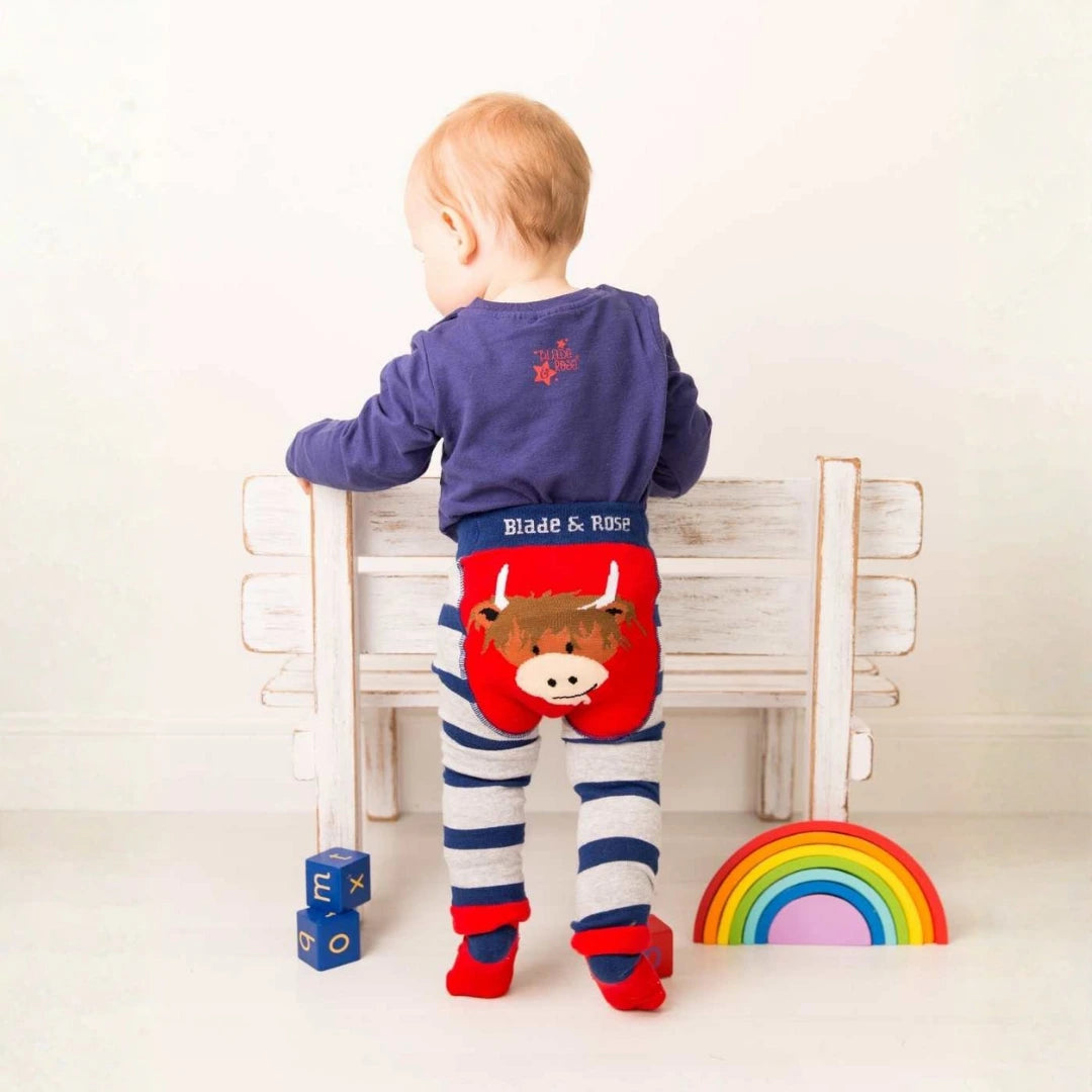 A baby standing on a wooden bench, viewed from behind, wearing red cow-print pants, navy striped leggings, and a navy long-sleeve top.