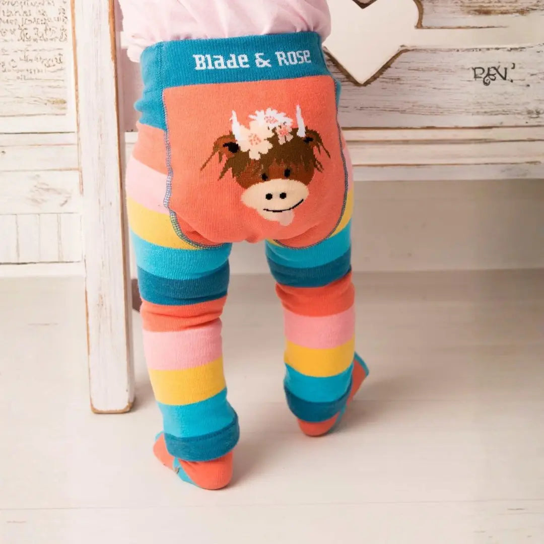 A child wearing colorful striped tights with a large cow patch on the back pocket. The tights feature horizontal stripes of blue, teal, pink, yellow, and orange, with a brown cow face and white flowers on the back pocket.