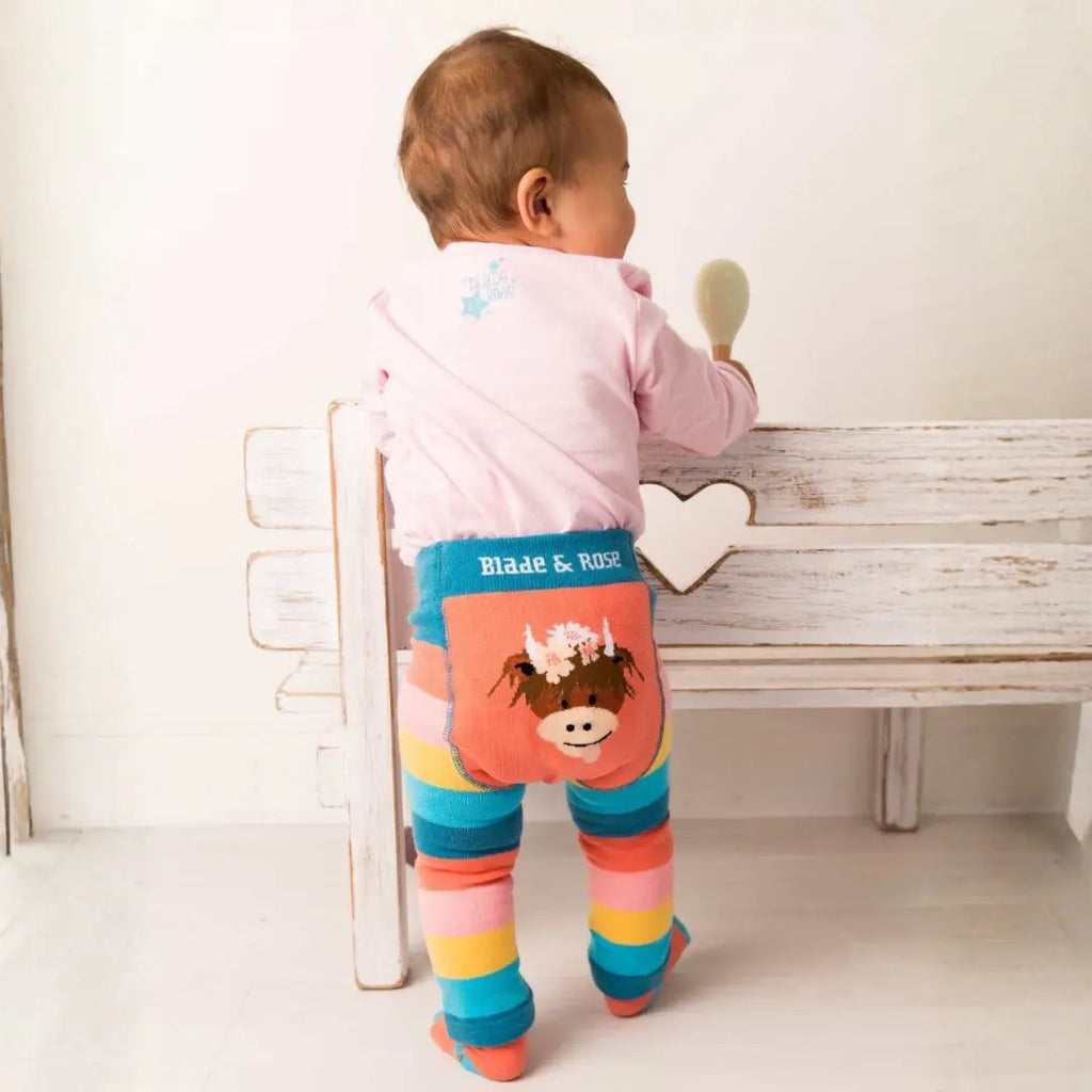A baby, back to camera, wearing striped tights with a cow applique and a pink top, next to a wooden bench.
