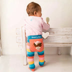 A baby, back to camera, wearing striped tights with a cow applique and a pink top, next to a wooden bench.