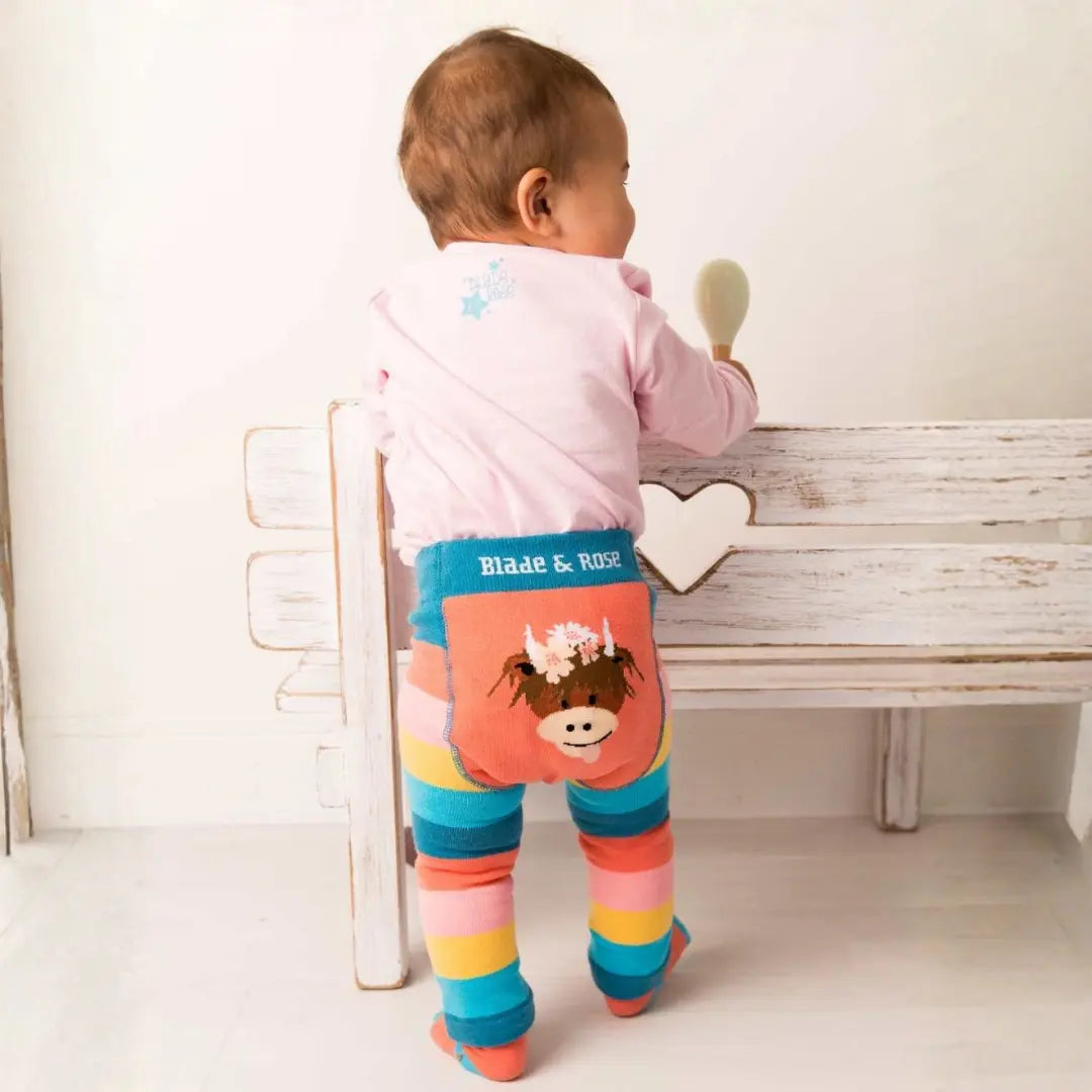 A baby standing with their back to the camera, wearing colorful striped tights with a cow applique and a pink top, positioned next to a rustic white wooden bench.