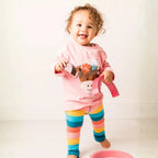 A smiling toddler wears a pink top with a cartoon graphic and striped leggings in yellow, blue, orange and pink, holding a toy watering can.