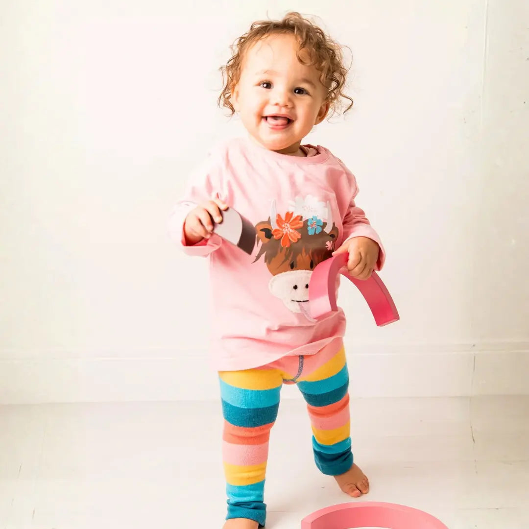A smiling toddler stands barefoot in a bright room, wearing a pink long-sleeve top with a cartoon character graphic and colorful striped leggings in yellow, blue, orange, and pink. The child is holding a pink toy watering can.