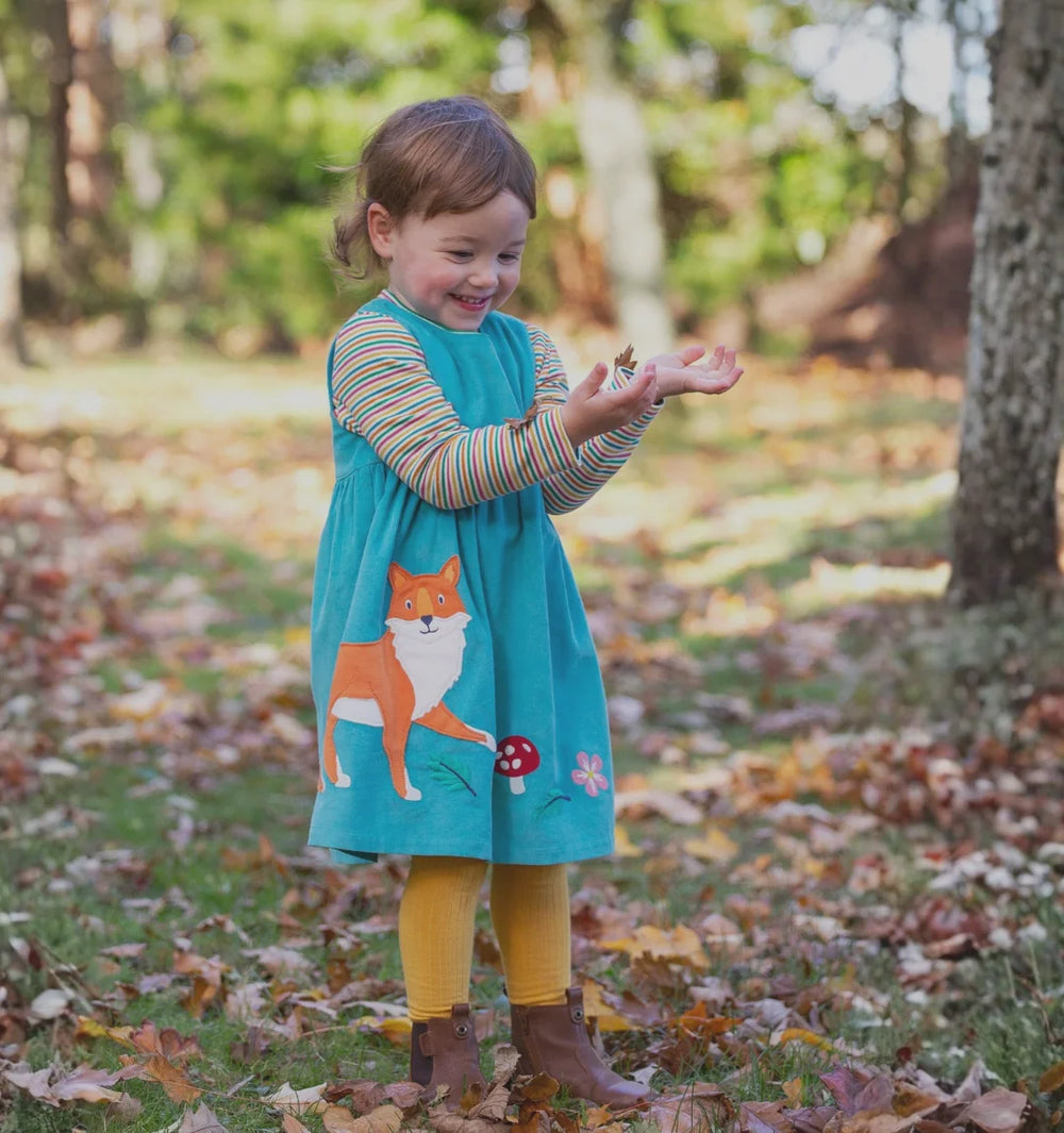 Child in a turquoise dress with fox design standing in a park with fallen leaves.