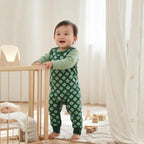 Baby in green pajamas standing next to a crib in a bright room.