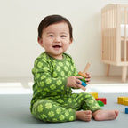 Baby in green pajamas with clover pattern sitting on a light blue mat in a room with a crib.