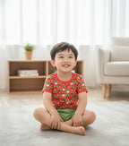 Child sitting on a rug in a living room wearing a red shirt with green shorts.
