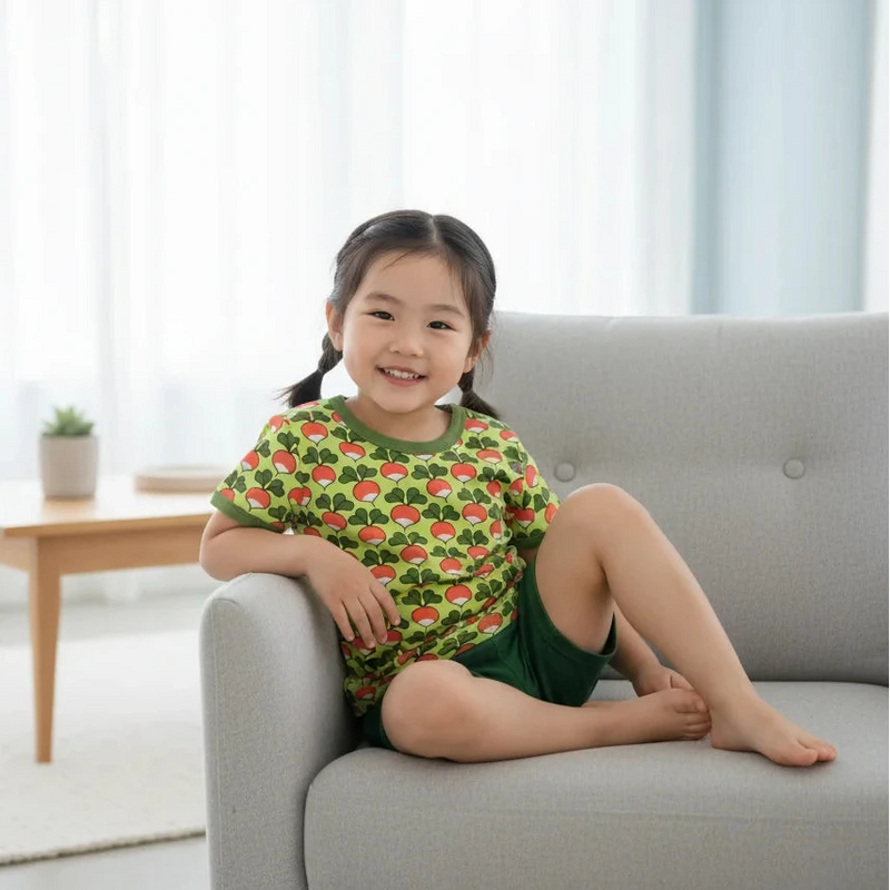Young girl sitting on a couch wearing a green shirt with red patterns and green shorts.