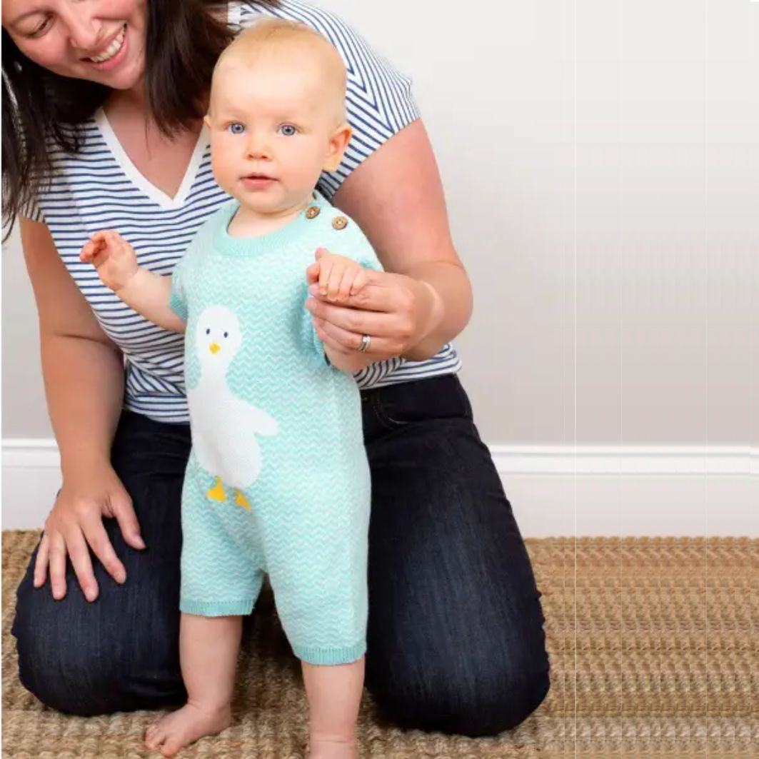Baby in a light blue outfit with a duck design, standing next to a person on a carpeted floor.