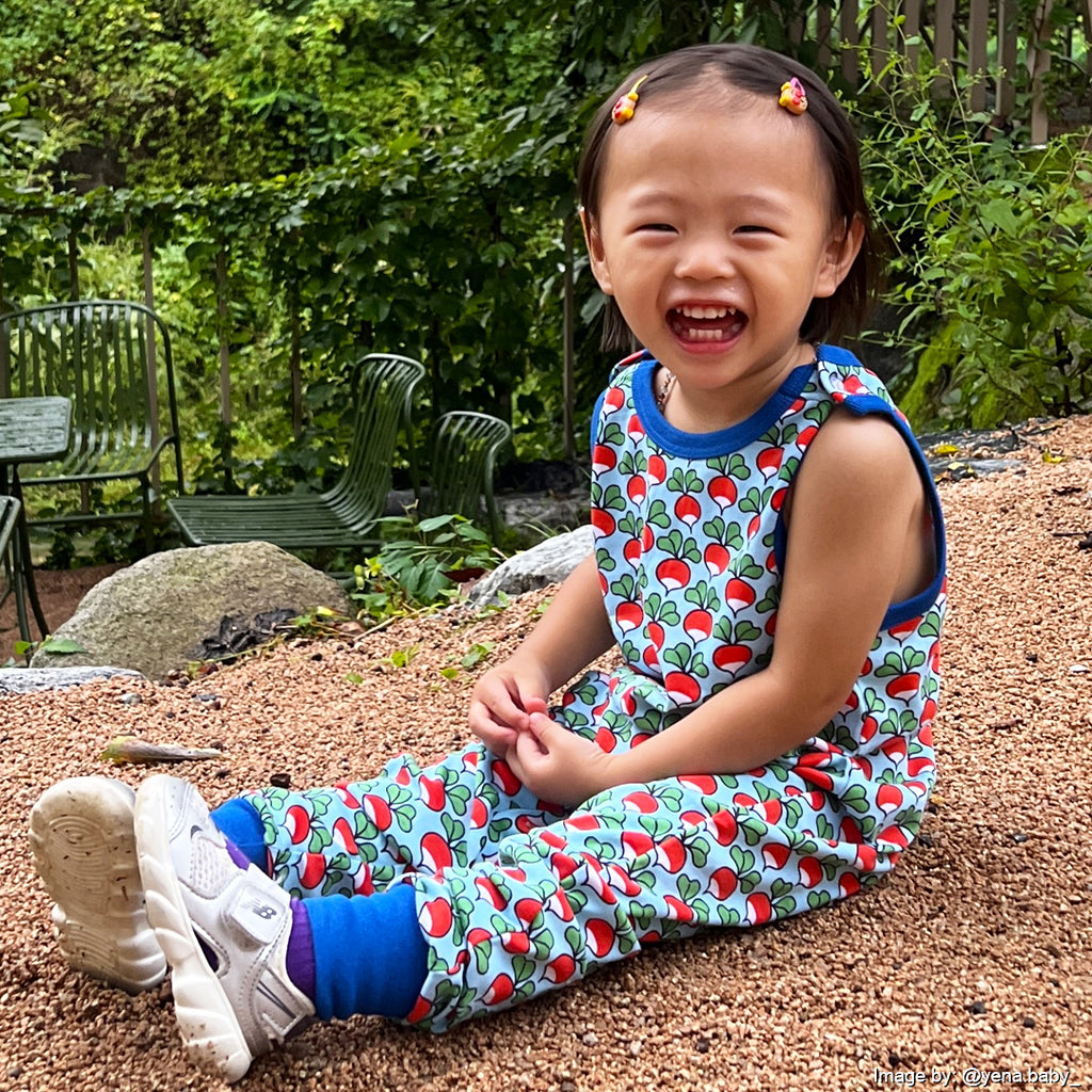 Child wearing a colorful outfit with a Radish pattern, sitting on a gravel path outdoors.