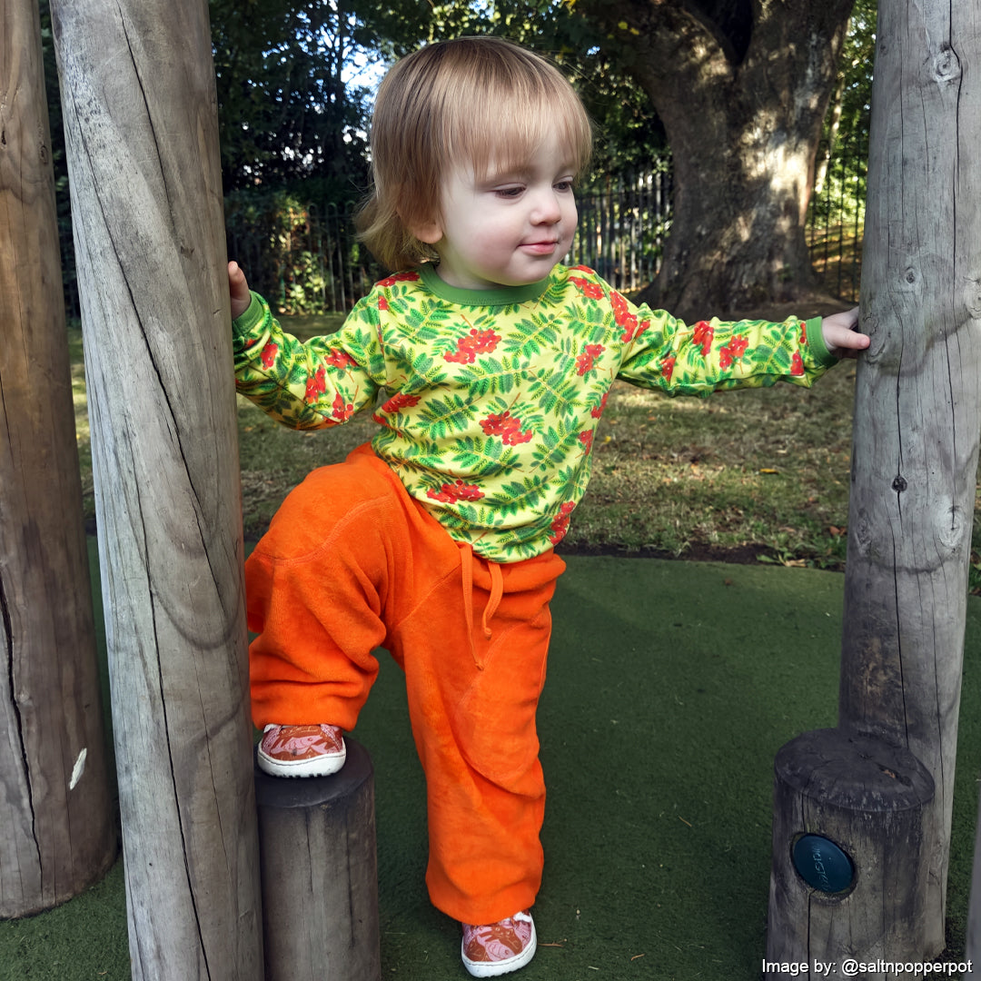 Child in a colorful shirt and orange pants standing between wooden posts outdoors.