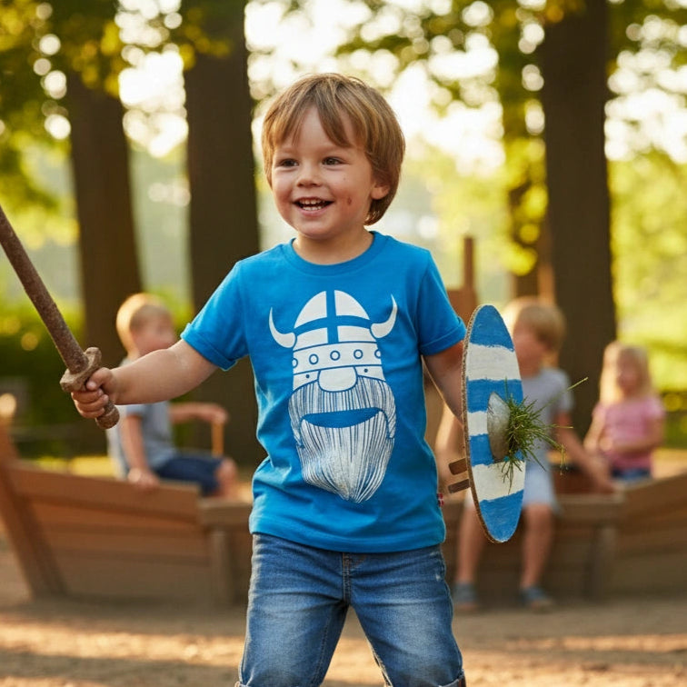 Child in a blue t-shirt with a beard graphic, holding a sword and shield, standing in a park.