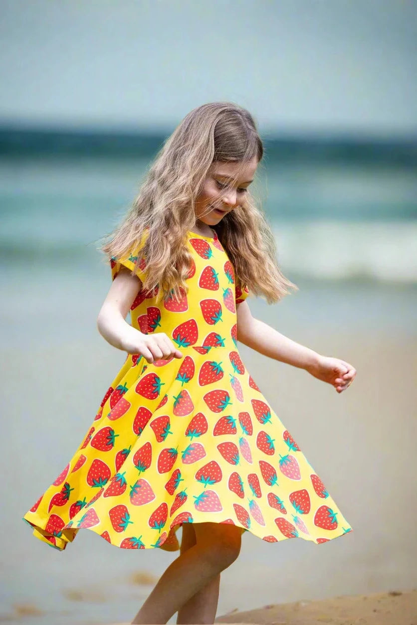 Young girl in a yellow dress with strawberry pattern standing outdoors in a park.