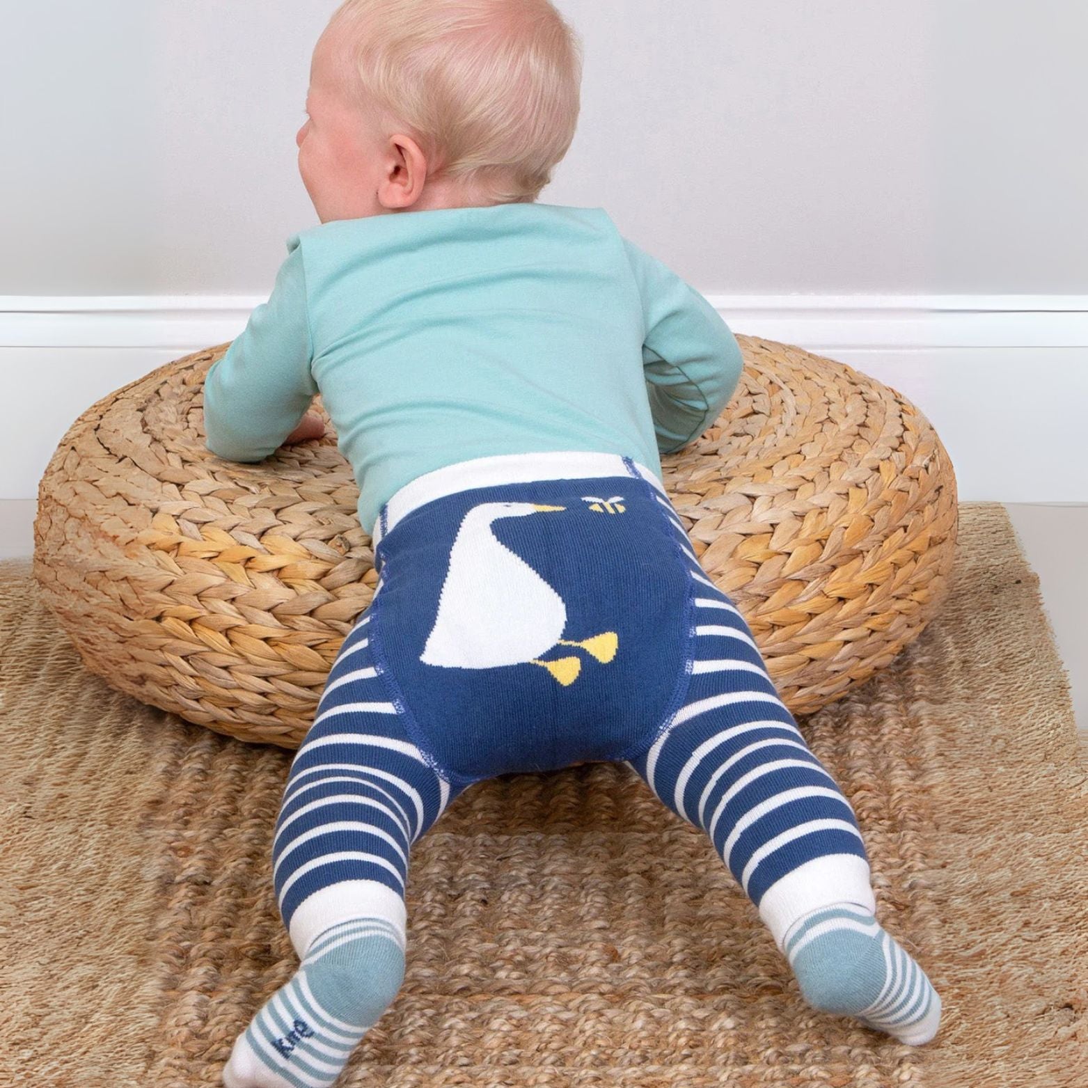 Baby wearing blue and white striped leggings with a duck design, standing on a woven mat.