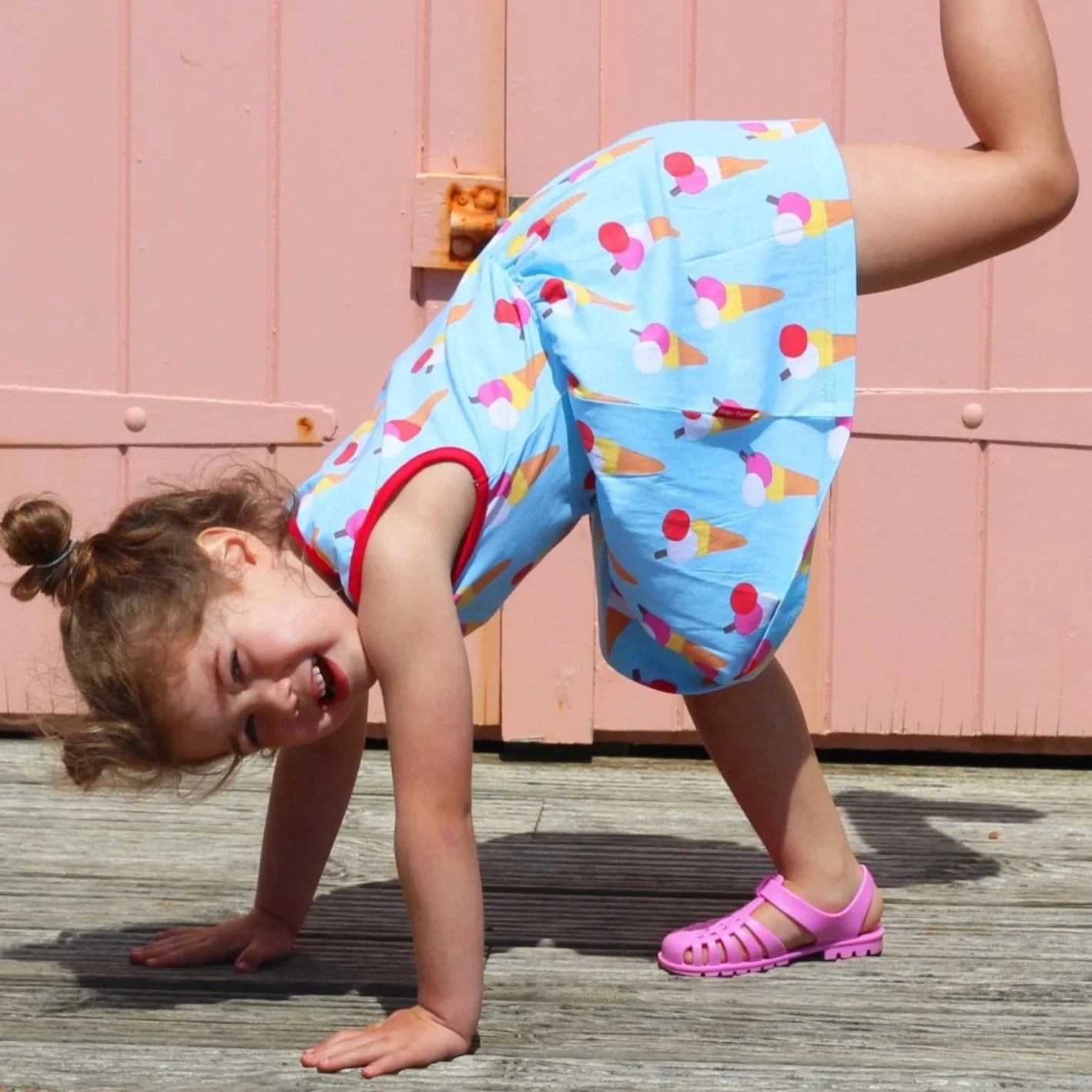Child in colorful ice cream-themed outfit doing a handstand on a wooden deck with a pink wall in the background.