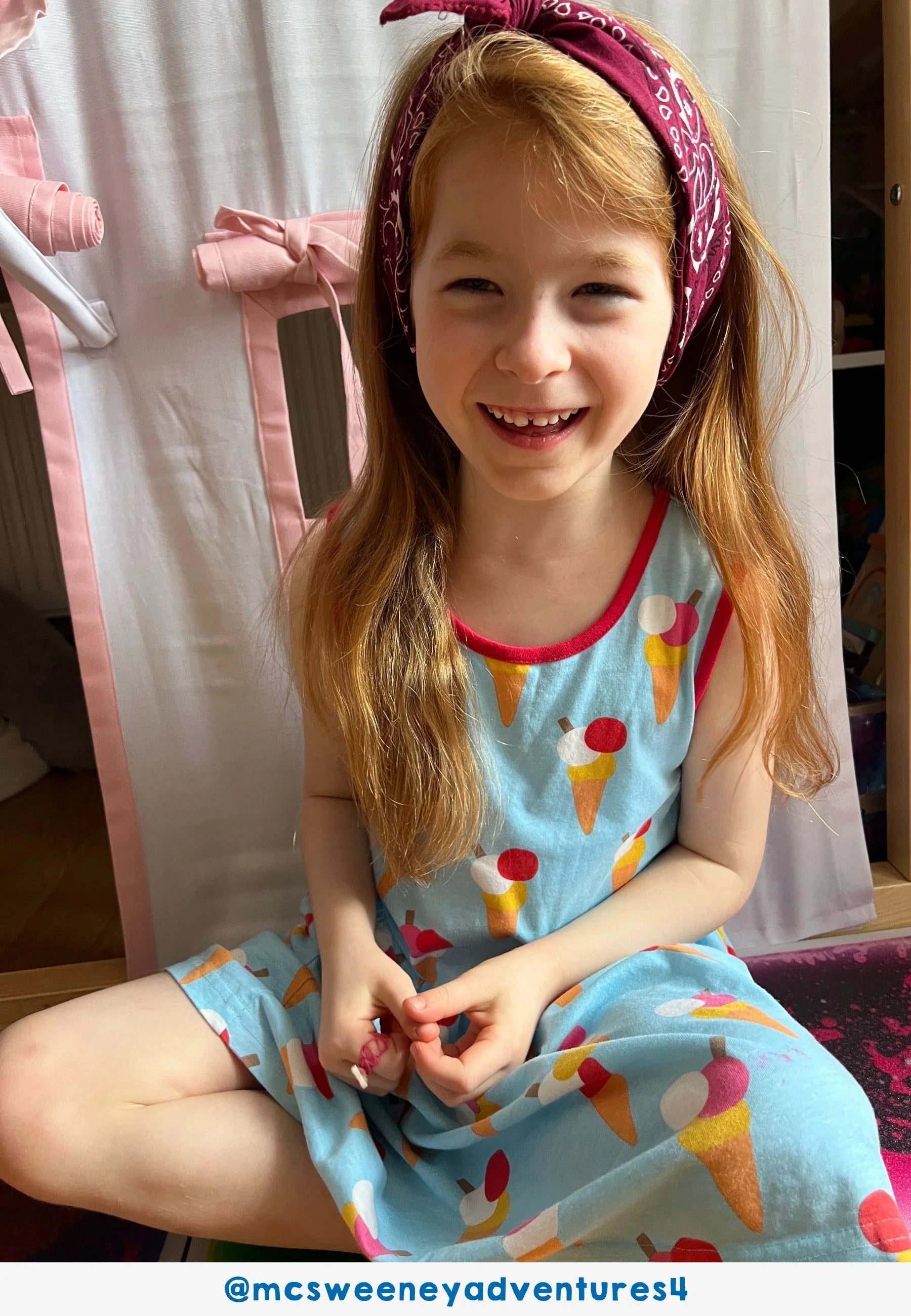 Young girl wearing a blue dress with ice cream cone pattern, sitting indoors.