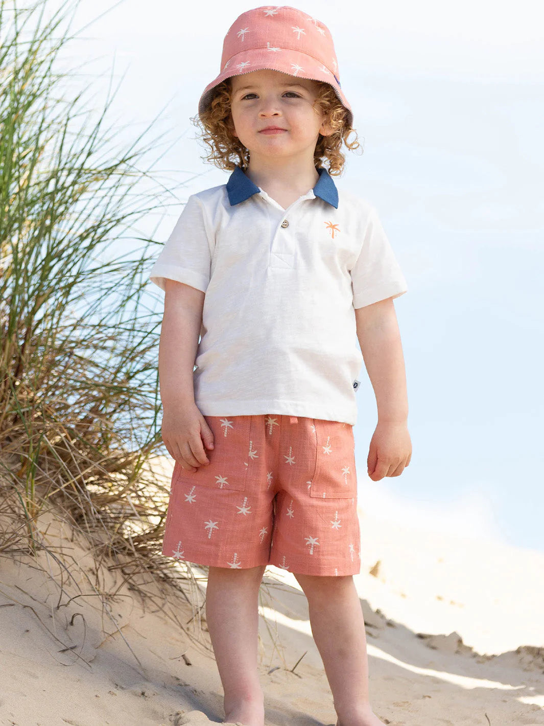Child wearing a white shirt with blue collar, pink shorts, and a pink sun hat on a sandy beach.