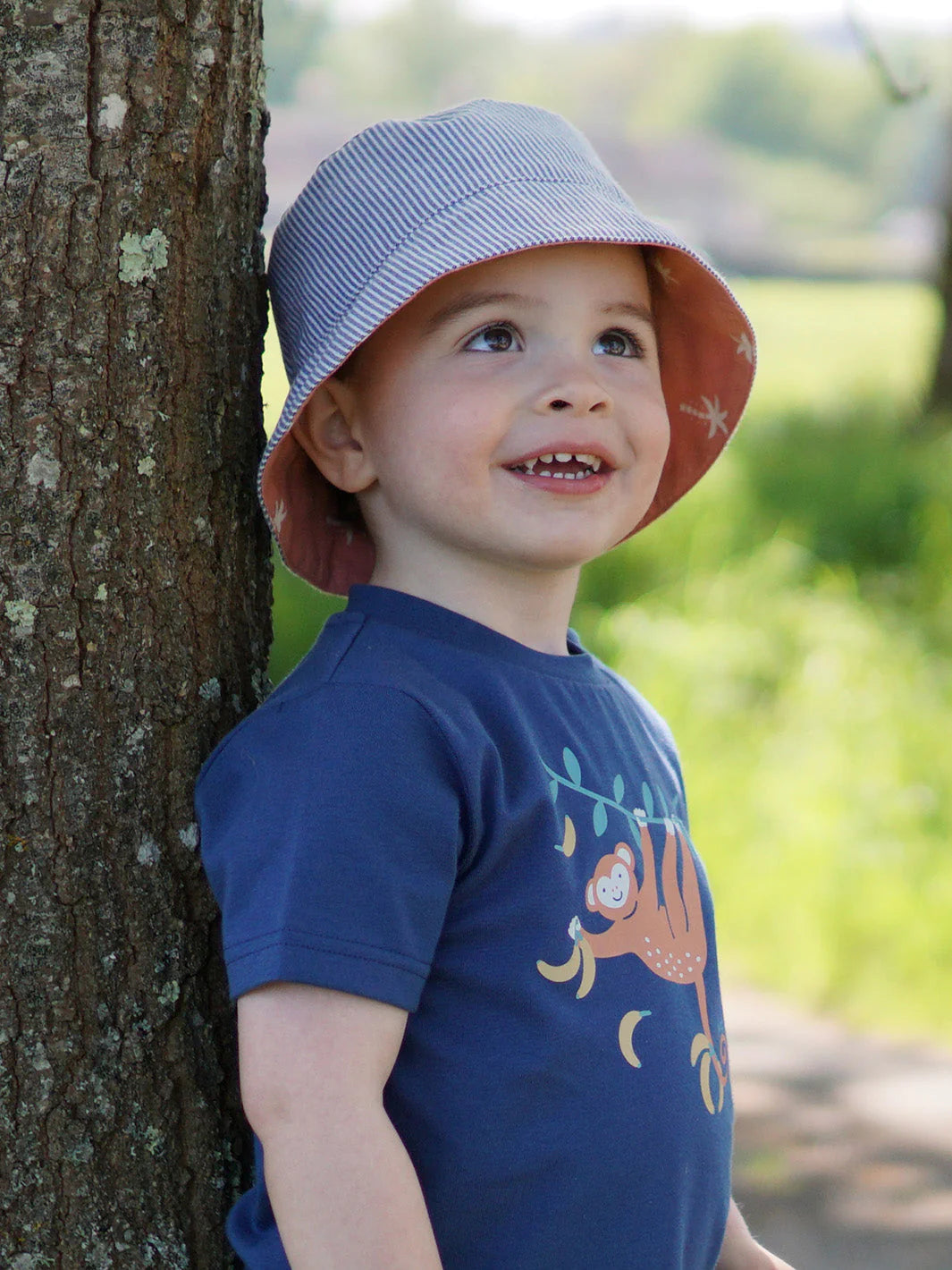 Child wearing a blue shirt with a graphic design, standing next to a tree in a natural setting.