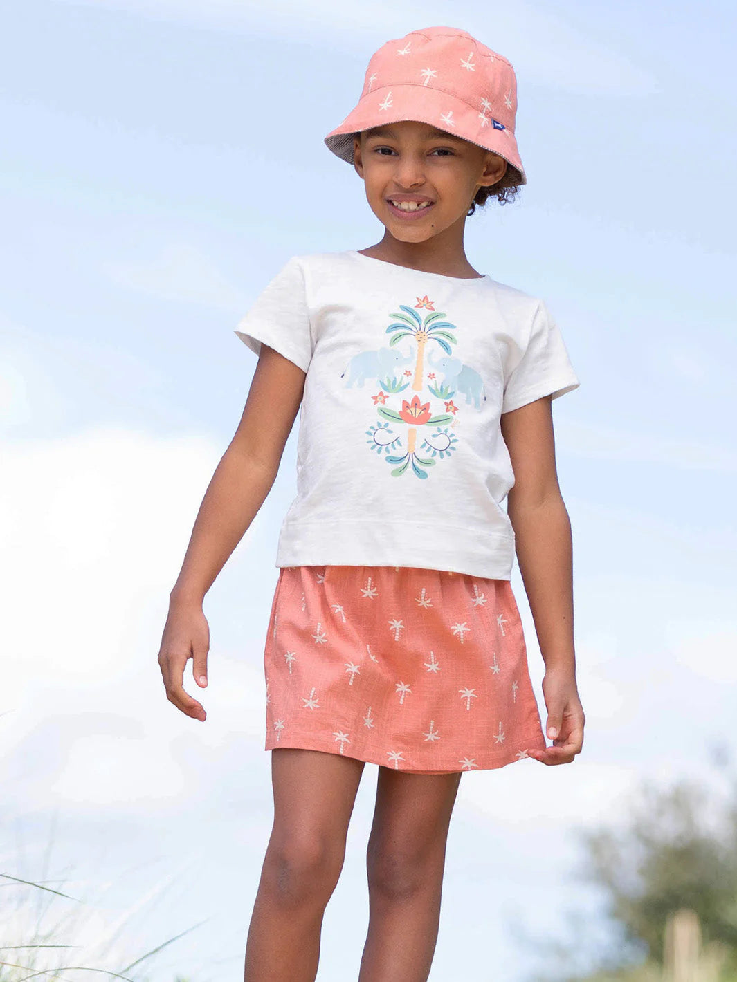 Young girl wearing a white t-shirt with a design, pink skirt, and pink hat outdoors.