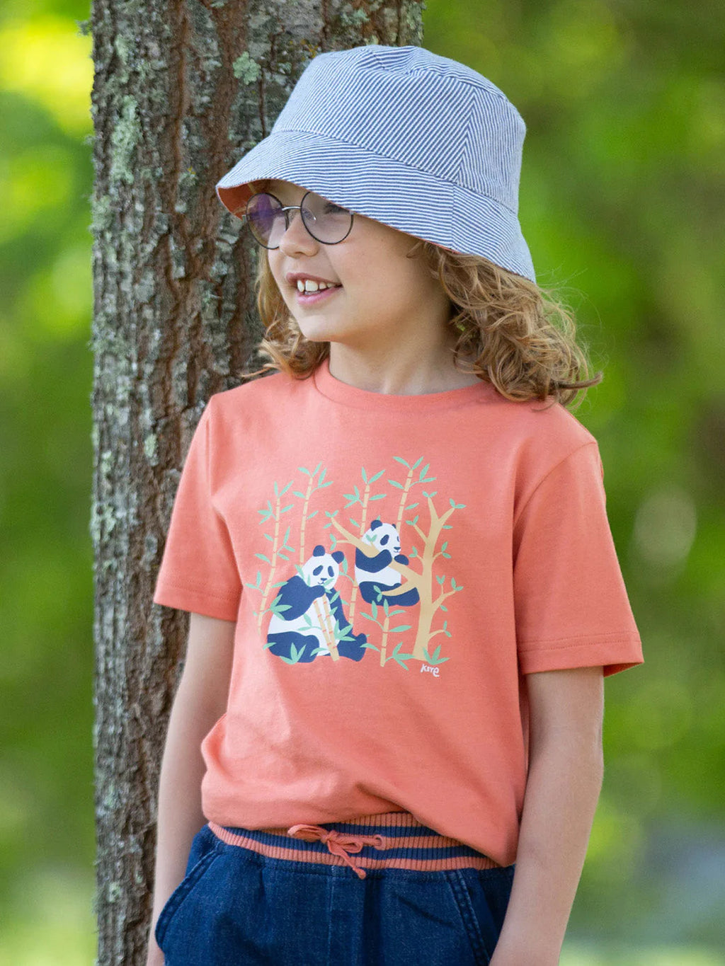 Child wearing a peach-colored t-shirt with panda design, blue shorts, and a light blue bucket hat, standing next to a tree.