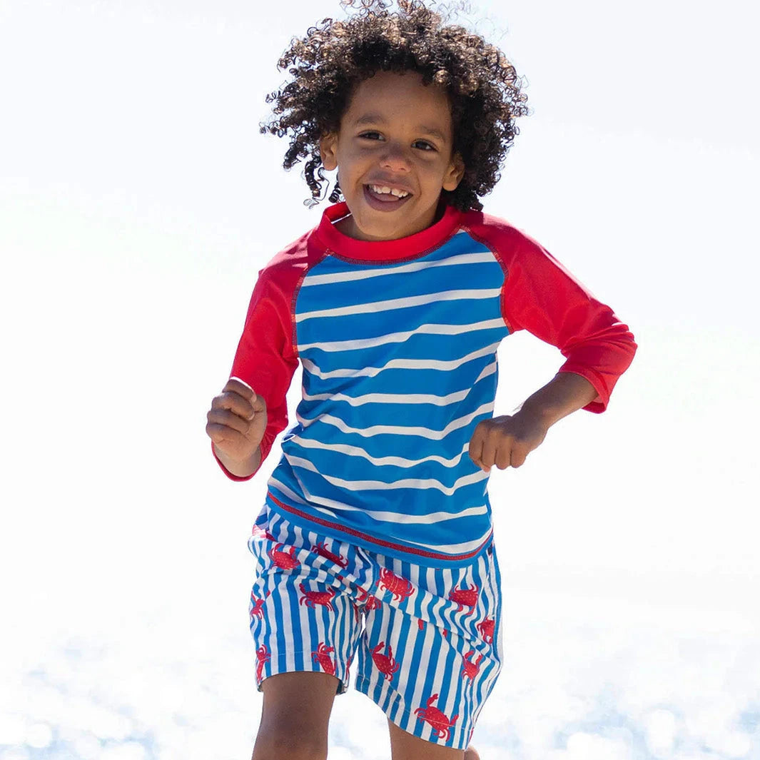 Child running on a beach wearing a blue and red striped swimsuit with a red shirt.