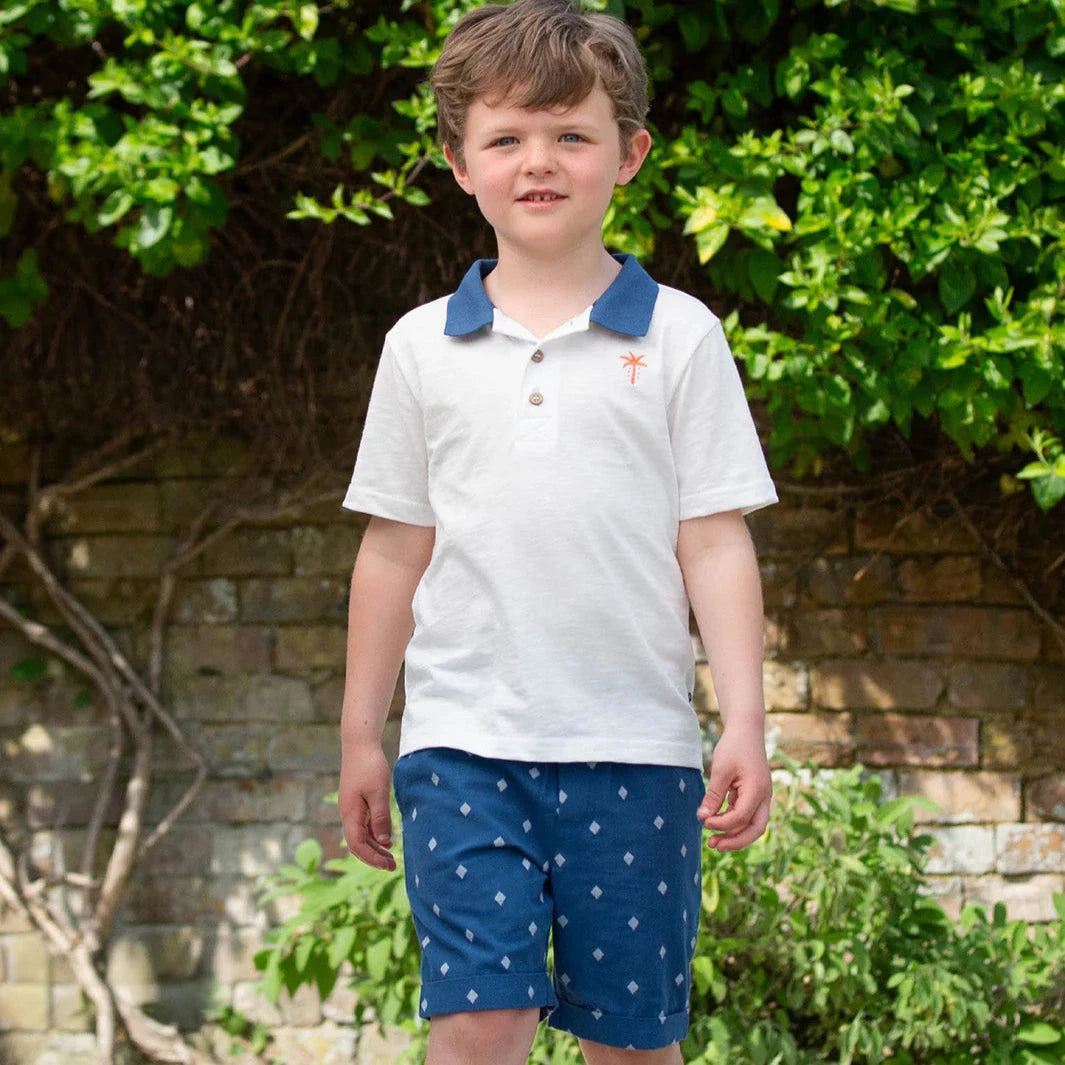 Young boy wearing a white polo shirt with blue accents and blue shorts with white patterns, standing outdoors.