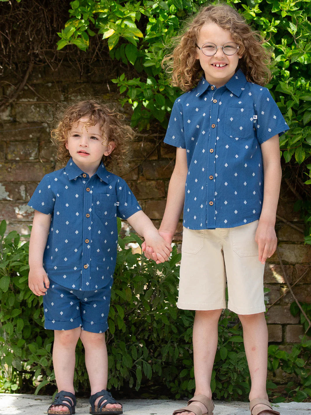 Two children wearing matching blue shirts with white patterns and beige shorts, standing outdoors.