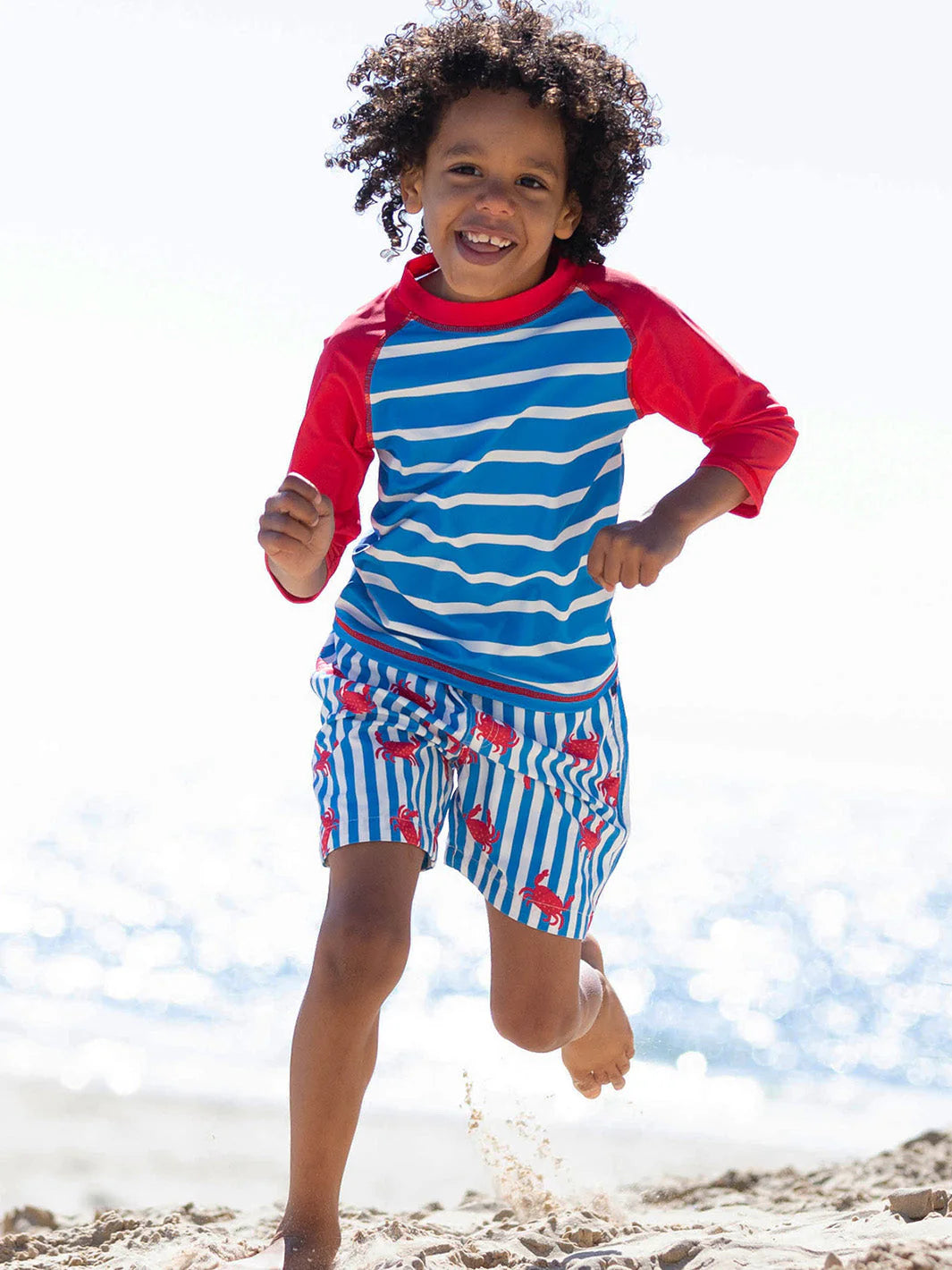Child running on a beach wearing a blue and red striped swimsuit with a red shirt.