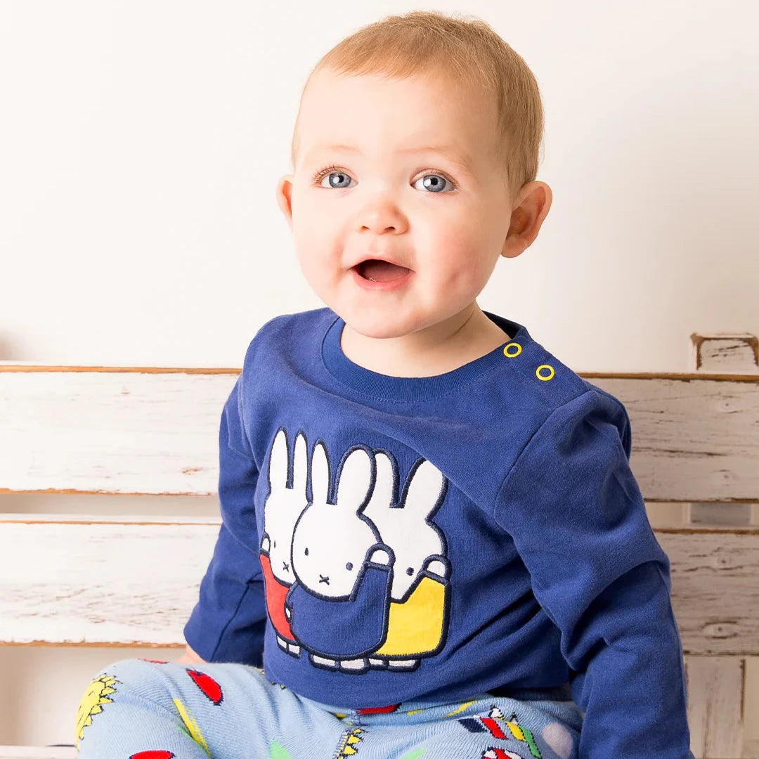 Baby wearing a blue shirt with a cartoon design and colorful pants sitting on a wooden bench.