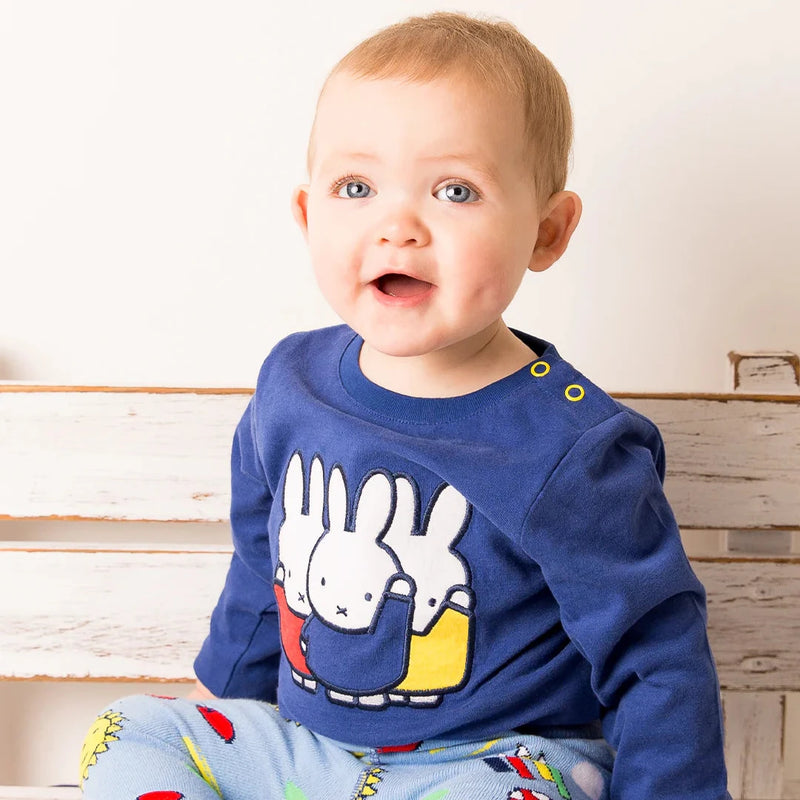 Baby wearing a blue shirt with a cartoon design and colorful pants sitting on a wooden bench.